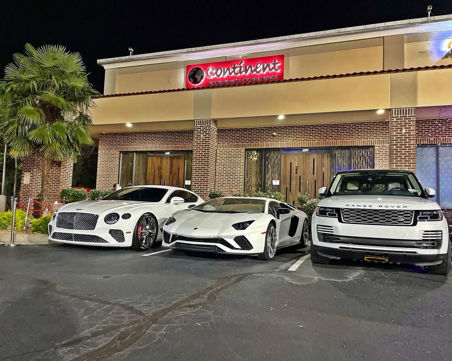 Three luxury cars parked outside a lit restaurant at night