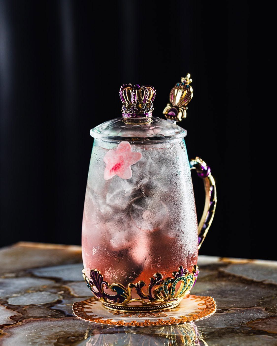 Pink fizzy drink in a jeweled glass mug with ice, on a dark background
