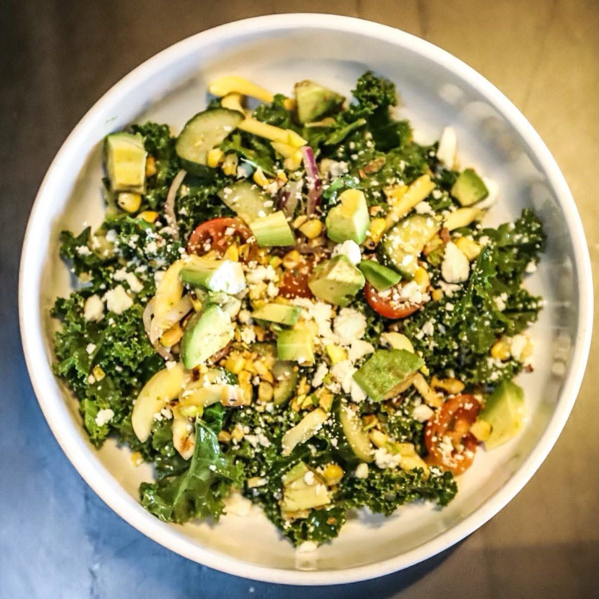 Bowl of kale salad with avocado, tomatoes, and seeds on a light surface