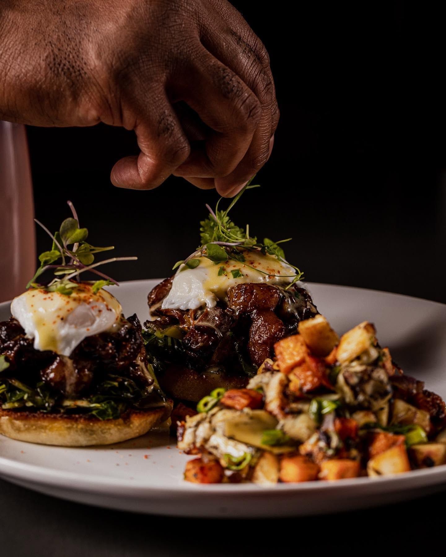 Hand placing herbs on two plated gourmet dishes beside a salad on a white plate