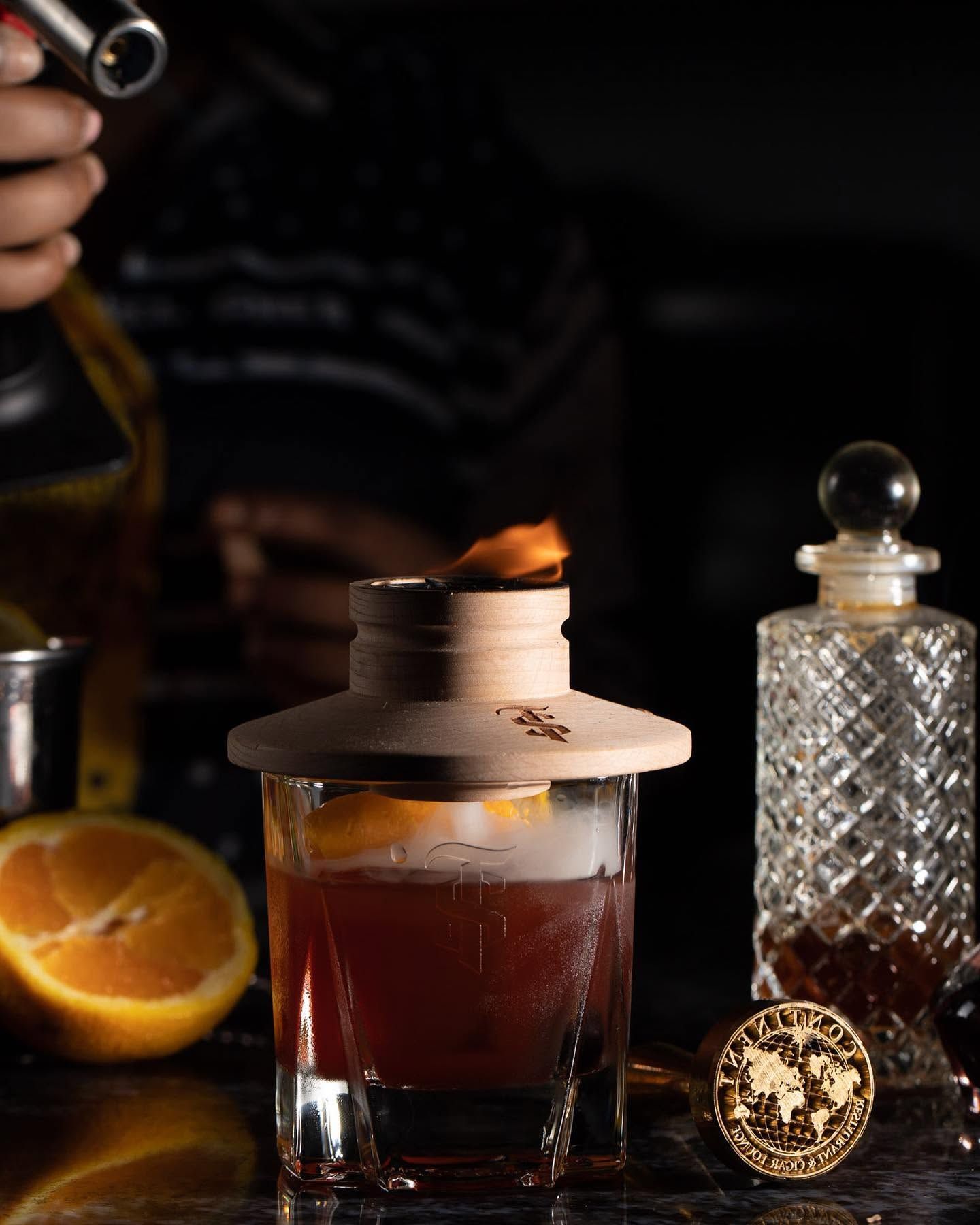 Cocktail on a dark bar with orange garnish, crystal decanter, and a lit match on top