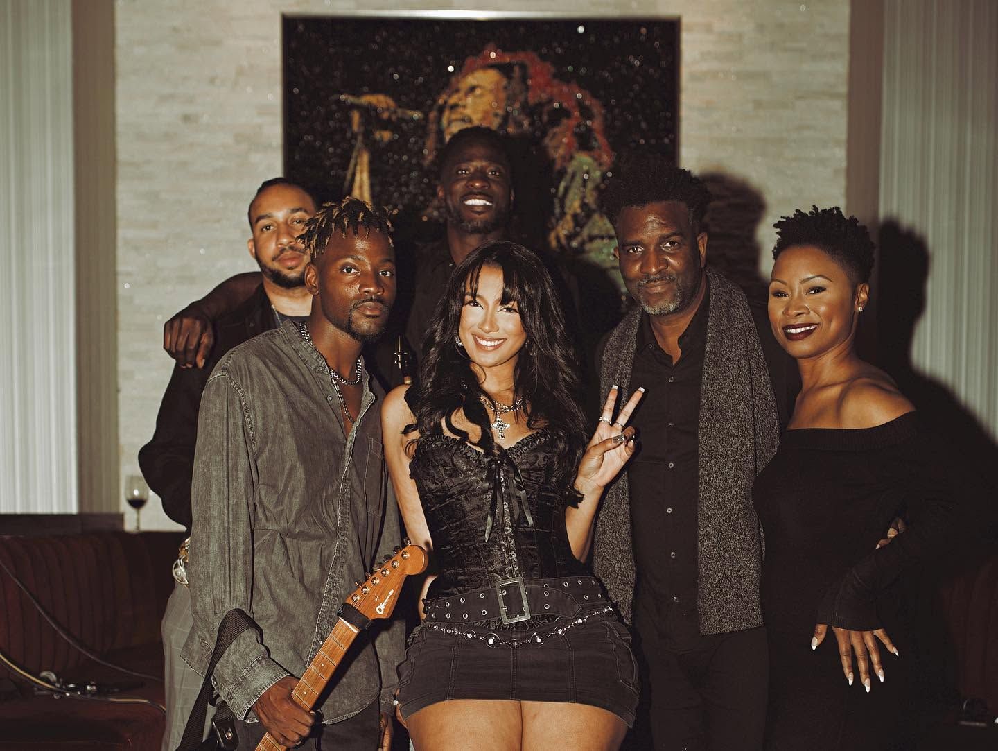 Group posing indoors at a party, with one woman making a peace sign and holding a guitar.