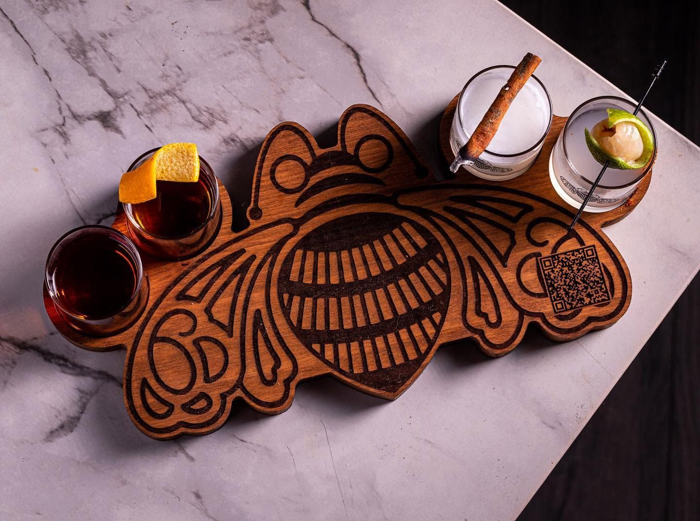 Decorative wooden serving tray with engraved floral pattern, surrounded by small glasses of drinks on a marble surface