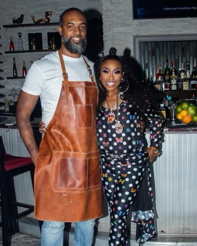 Two people posing in a bar, one in a leather apron and the other in a patterned outfit, smiling.
