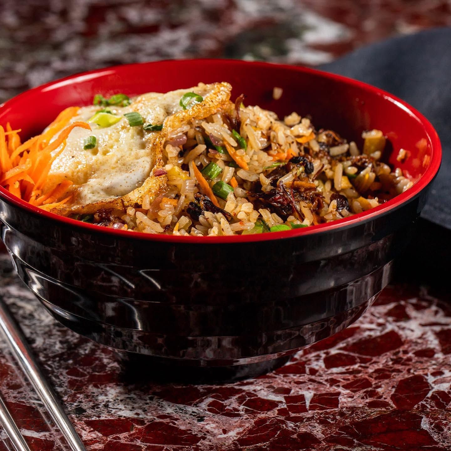 Red bowl of rice topped with vegetables and sauce on a marbled table