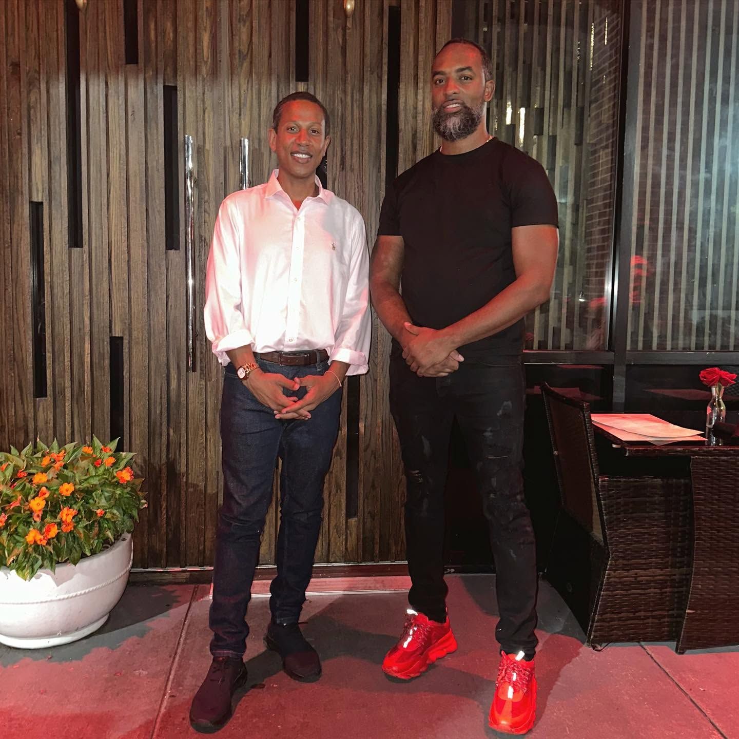 Two people stand indoors by a wood-paneled wall, one in a white shirt and jeans, the other in black with red sneakers.
