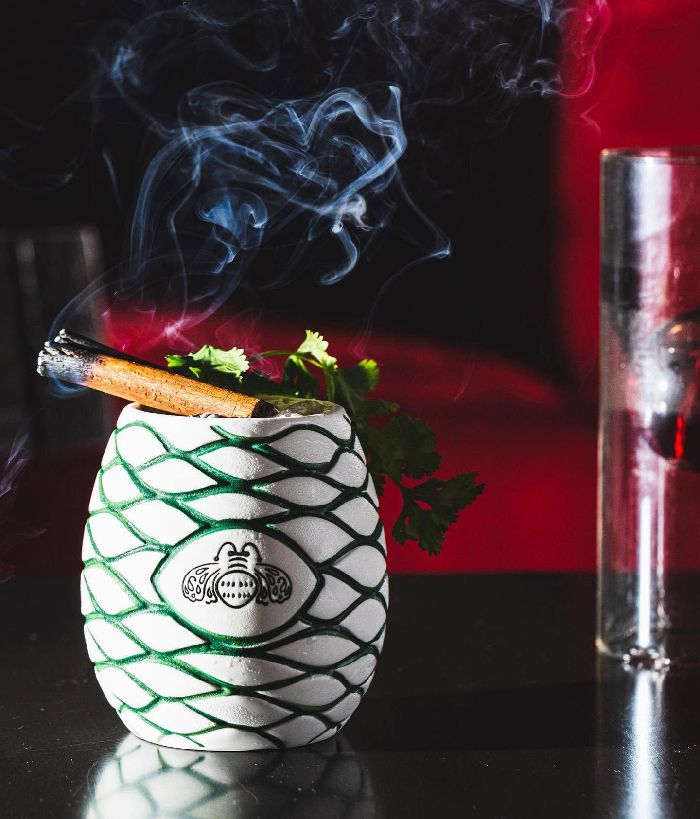 Smoking cocktail in white pineapple mug with green garnish and lime, on dark bar with red background