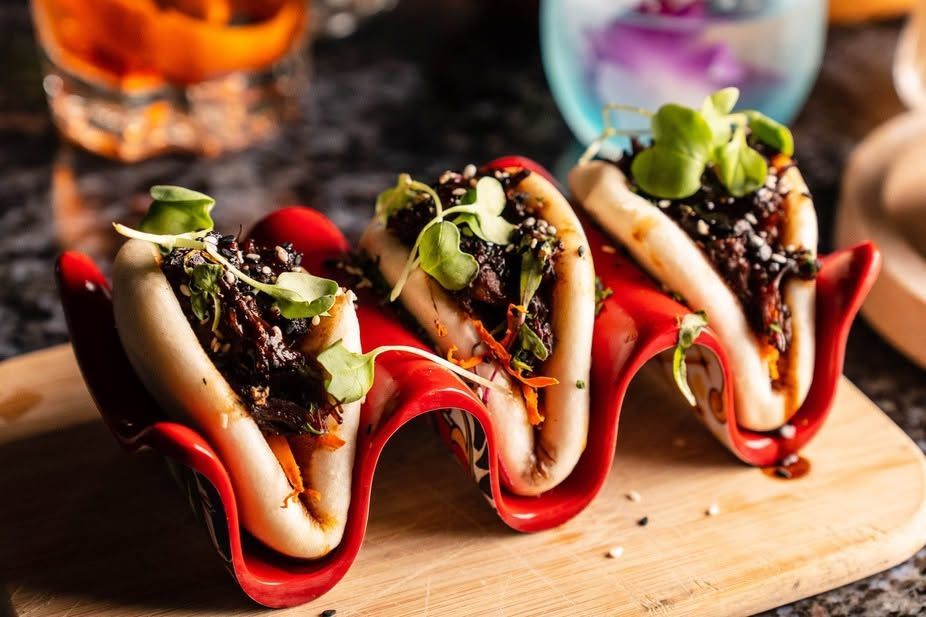 Three gourmet hot dogs with toppings on a wooden board, lit warmly and served with dipping sauce.