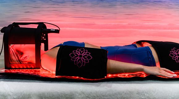 Person lying under red light therapy, black device and wraps, beach background.