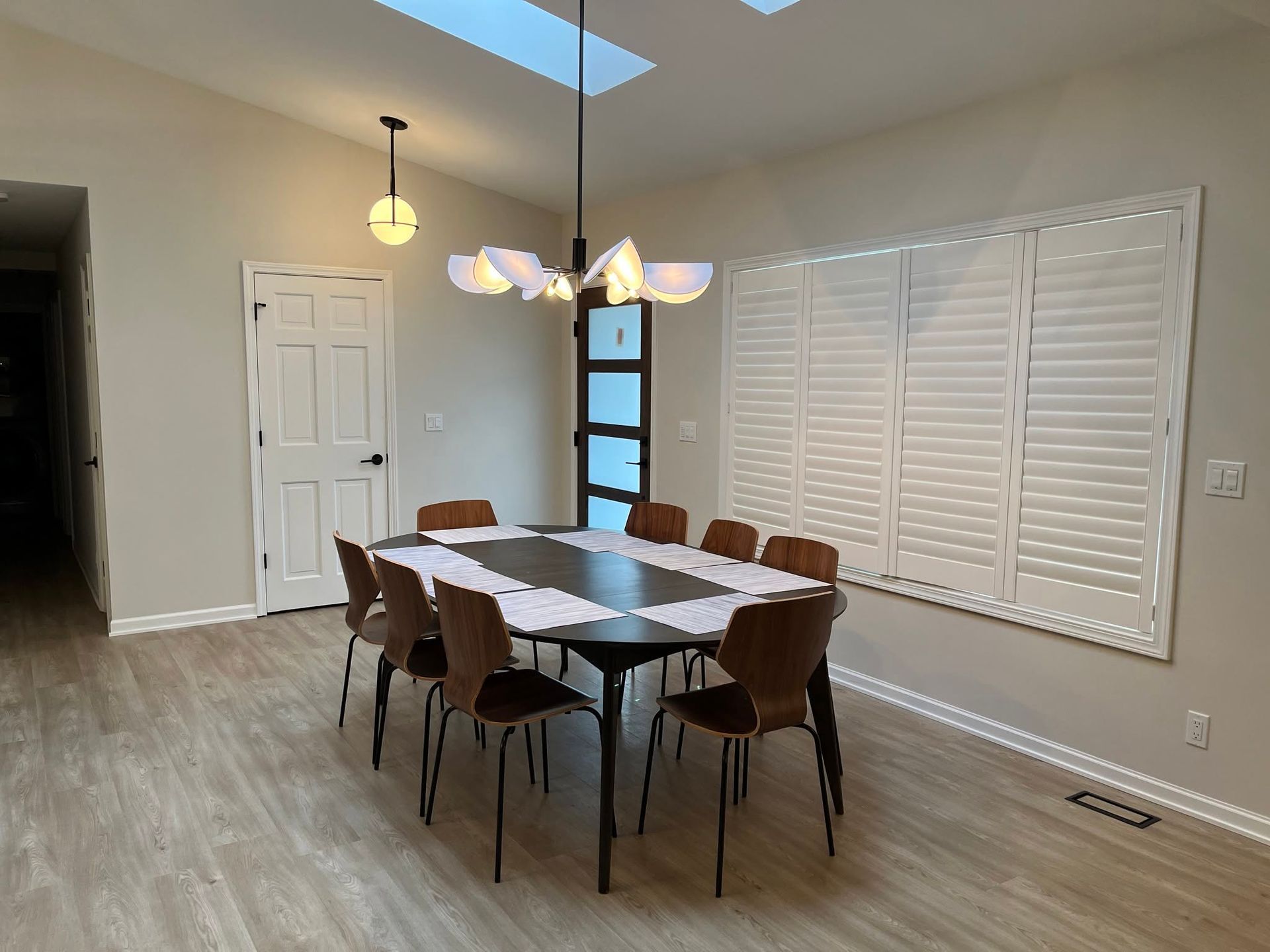 Dining room with a table and six chairs, pendant light, and large window shutters on a hardwood floor.