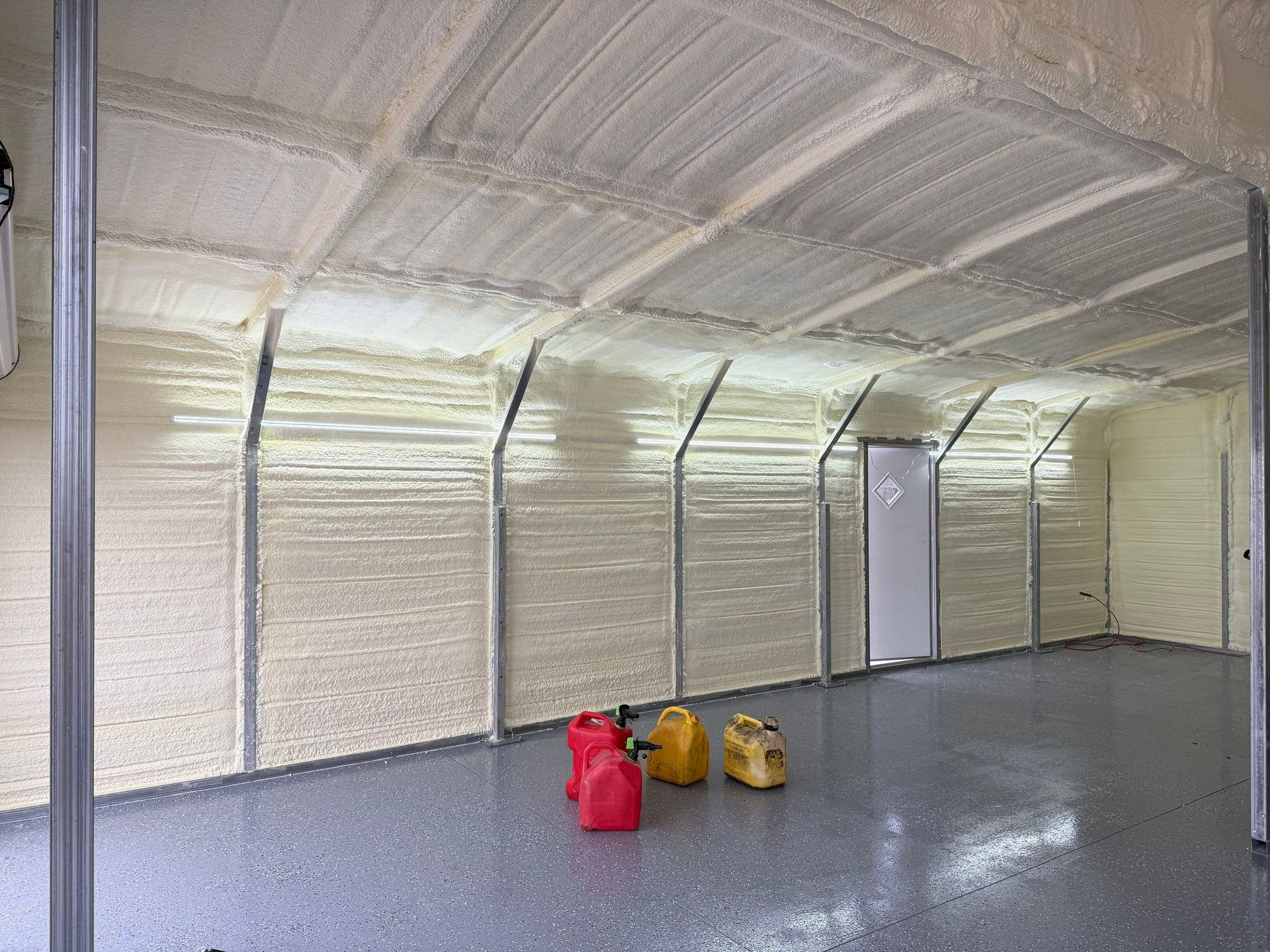 Insulated garage interior with light-colored spray foam on walls and ceiling, metal beams, and a gray epoxy floor.