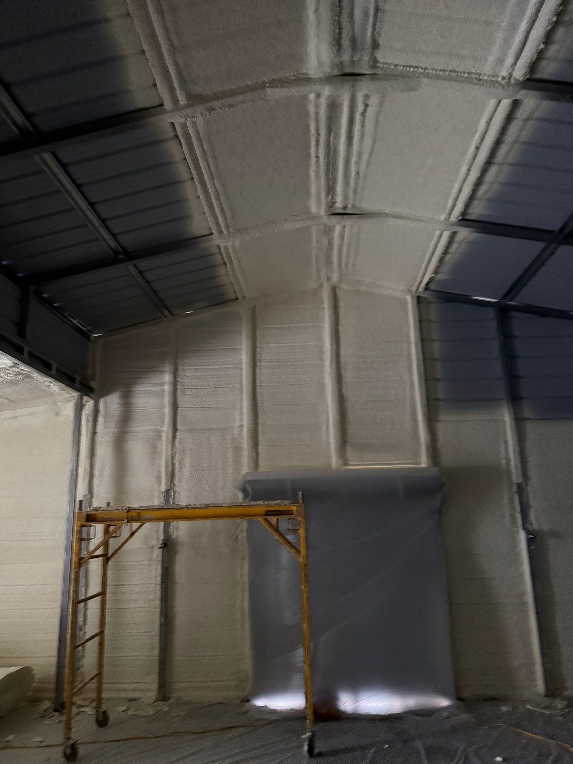 Interior of a metal building with spray foam insulation on the walls and ceiling. A yellow scaffold is in view.