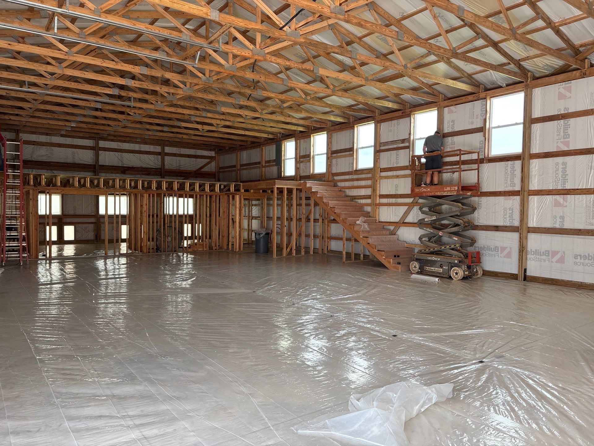 Interior of a large unfinished building with concrete floor, wooden frame, and a person on a lift near staircase.