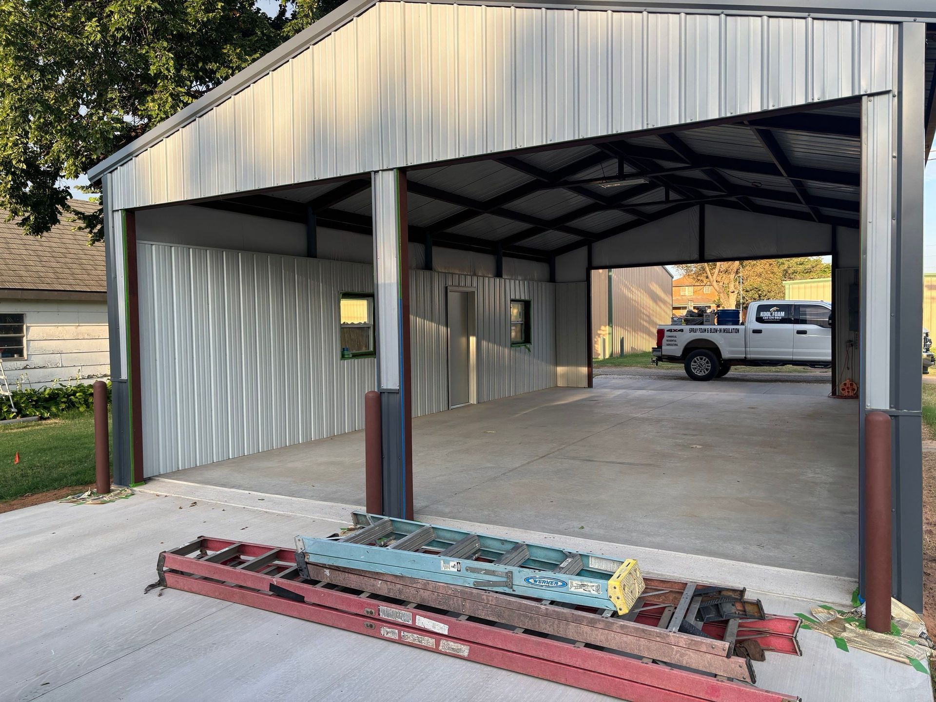 Metal-framed carport with a concrete floor, a truck parked inside. Construction materials are in the foreground.