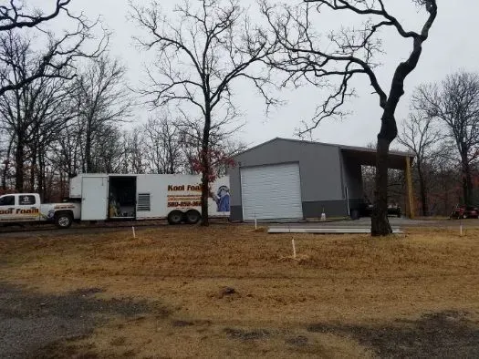 Truck and trailer parked by a gray metal building with a white garage door in a wooded area.