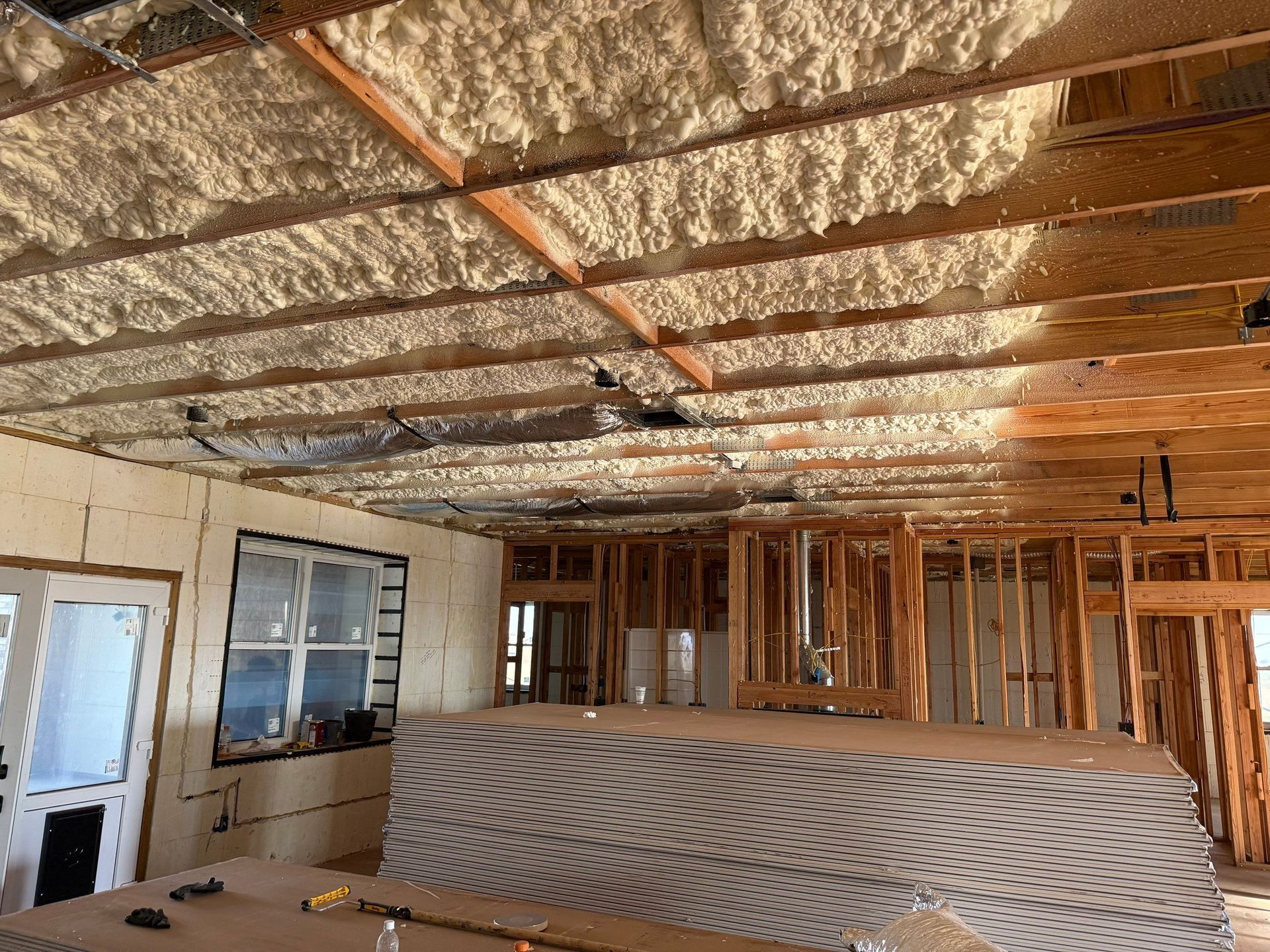 Interior view of a building under construction, spray foam insulation on ceiling, exposed studs, drywall stacked, and window.
