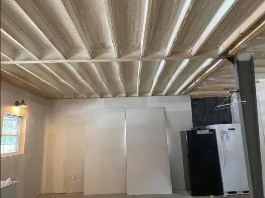 Basement ceiling with exposed beams, insulation, and drywall leaning against the wall. Refrigerator and window visible.