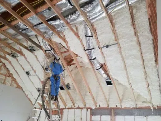Person spraying insulation foam between wooden rafters in a building.