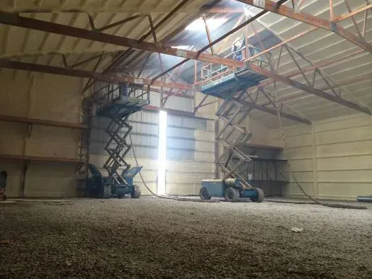 Interior view of a building being insulated with spray foam; workers on lifts. Light-colored walls and ceiling.
