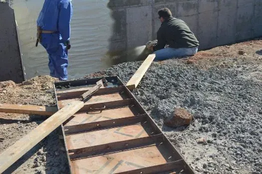 Workers near water, one in blue overalls, another seated. Metal frame and wood planks on gravel.