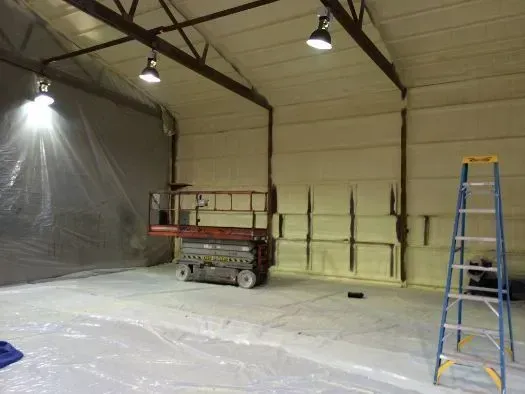 Interior of a building being insulated with foam.  A lift, ladder, and lights are present.