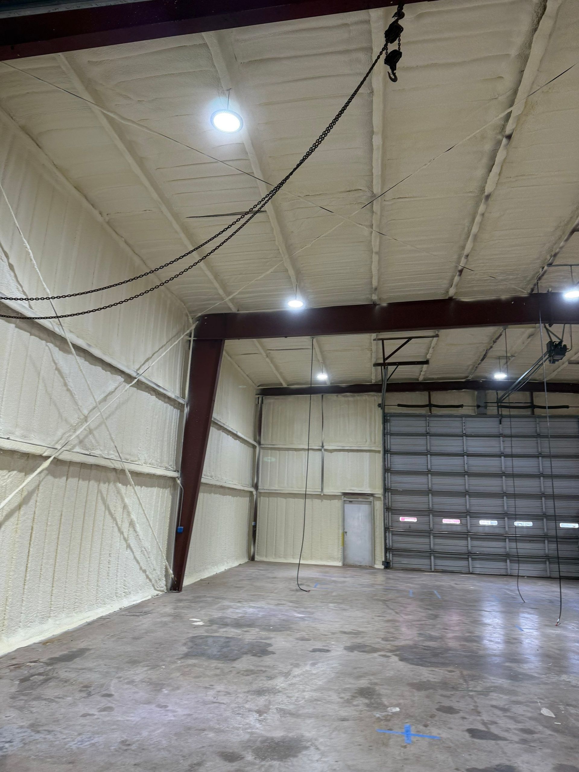 Interior of a warehouse with spray foam insulation, concrete floor, garage door, and metal beams.