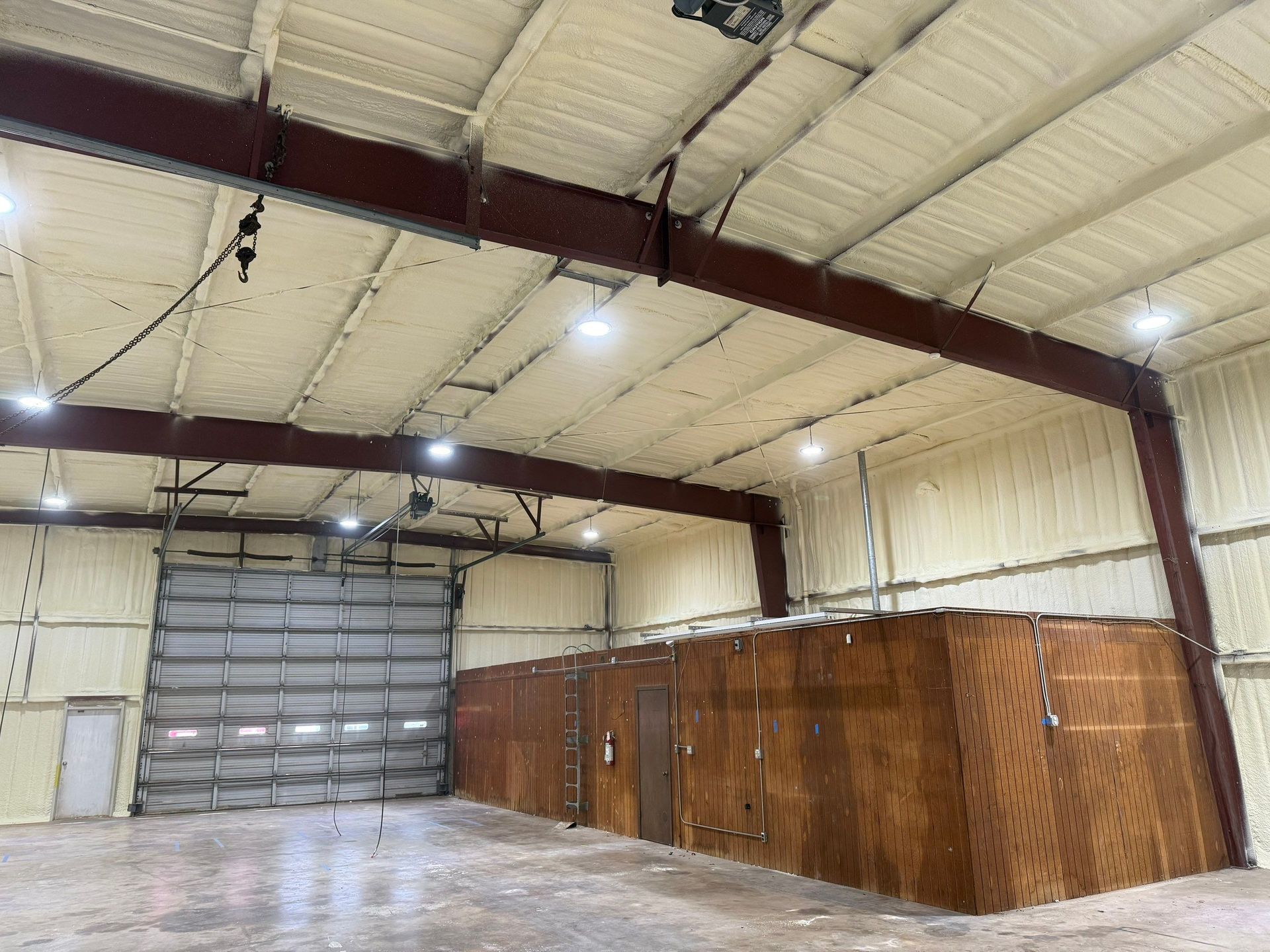 Inside a warehouse with spray foam insulation on ceiling and walls. Steel beams, overhead lights, large door, and wood paneling on a side wall.