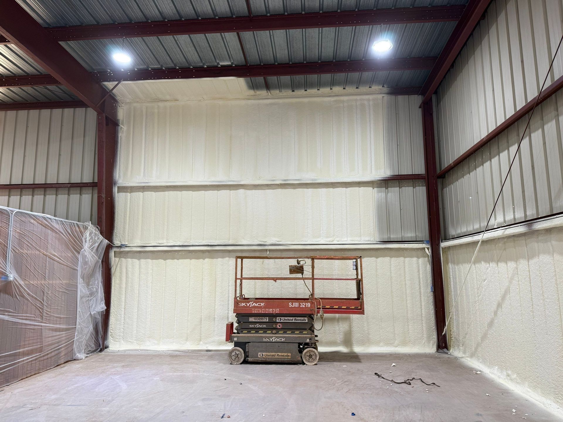 Interior view of a building with spray foam insulation on walls, red metal beams, and a lift.