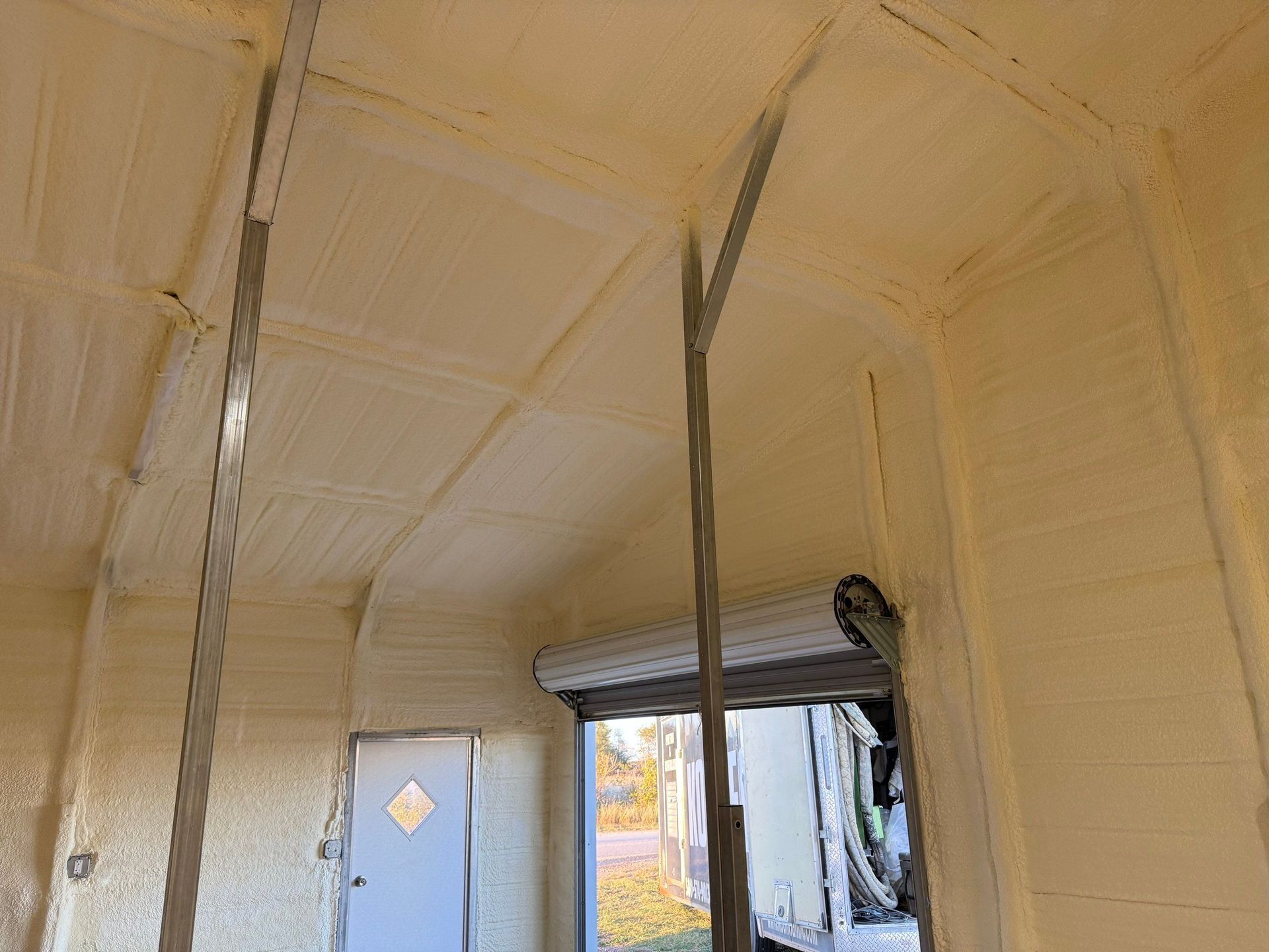 Interior view of a structure insulated with yellow foam, with metal beams, a door, and a partially open garage door.