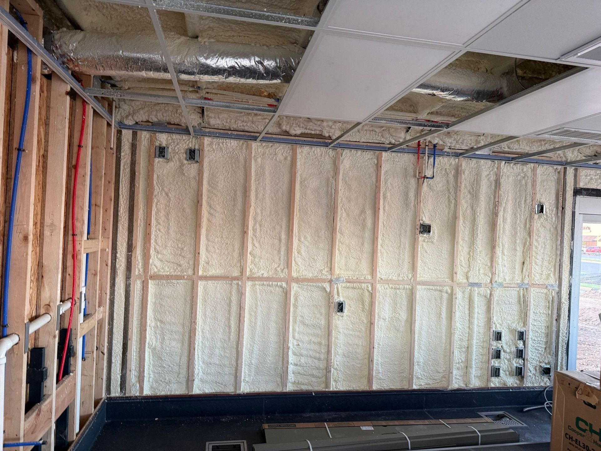 Interior of a building under construction, spray foam insulation applied to wooden wall studs, exposed piping, and ceiling grid.