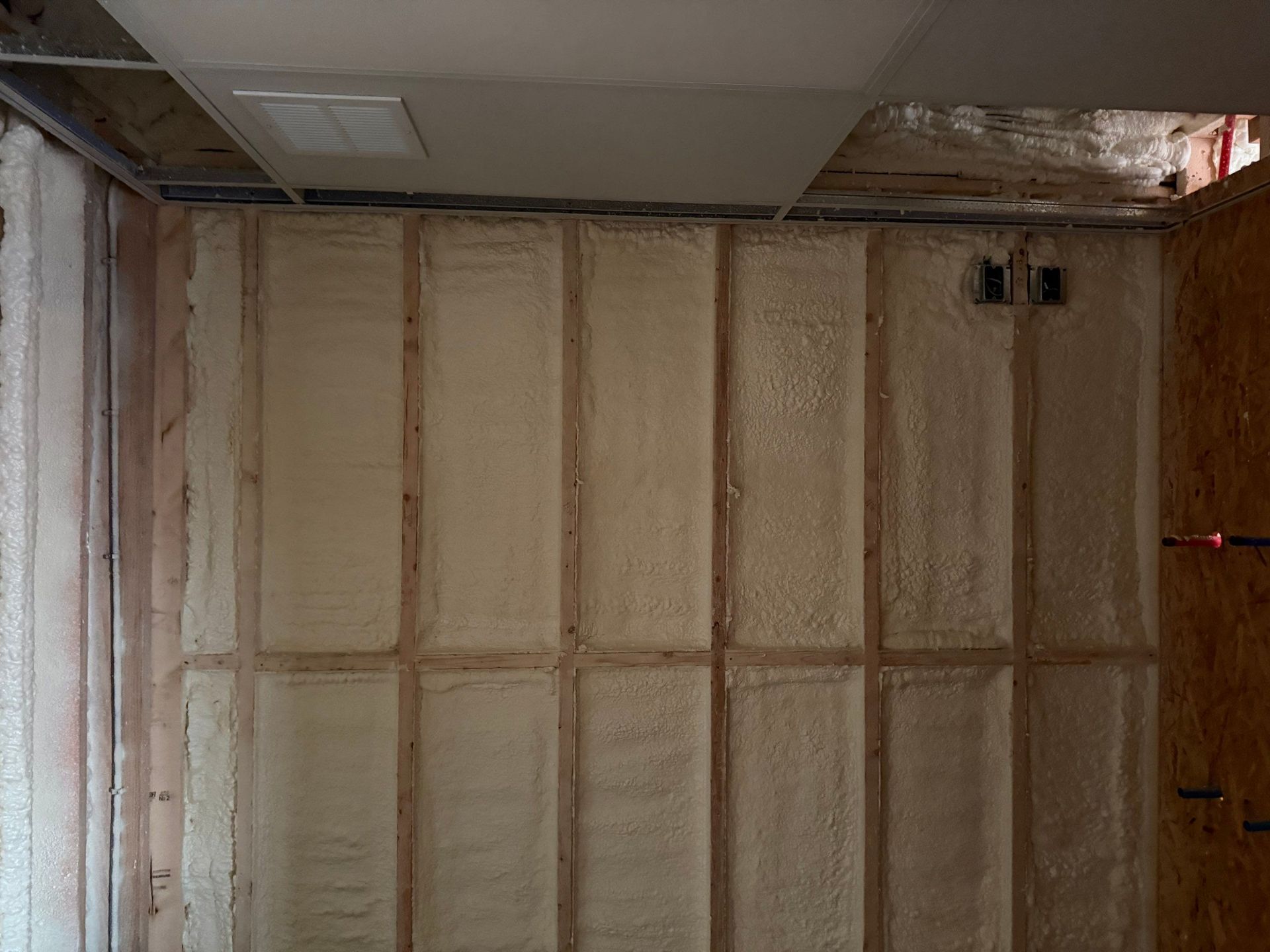 Interior wall with spray foam insulation between wooden studs, partially covered by drywall.