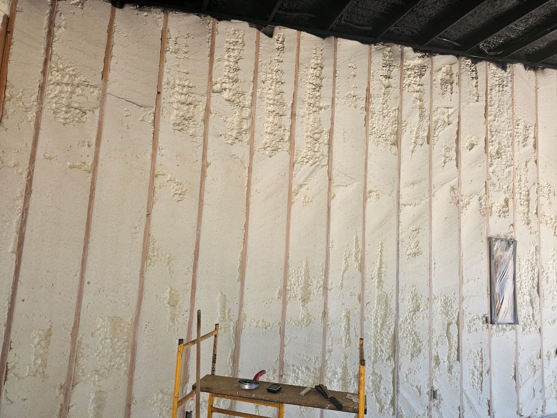 Wall insulation in progress with exposed wooden studs. Beige foam covers most of the wall.