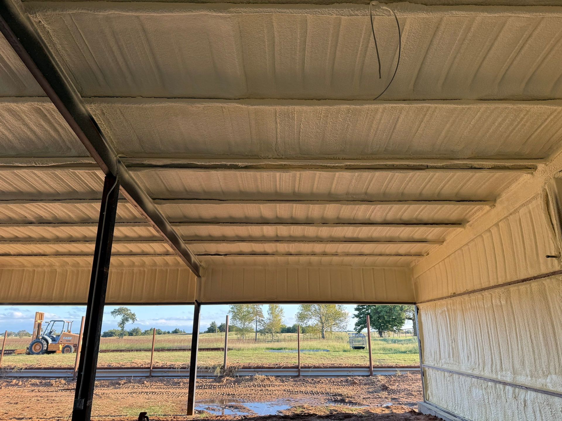 Interior view of a building with spray foam insulation on the roof and walls; exterior view of a field and a vehicle.