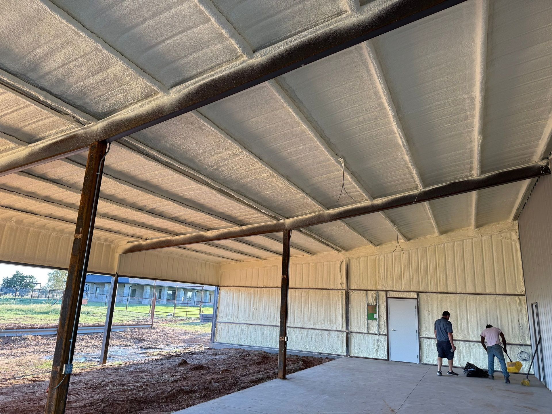 Interior of a building being insulated with spray foam. Two workers near a doorway, daylight outside.