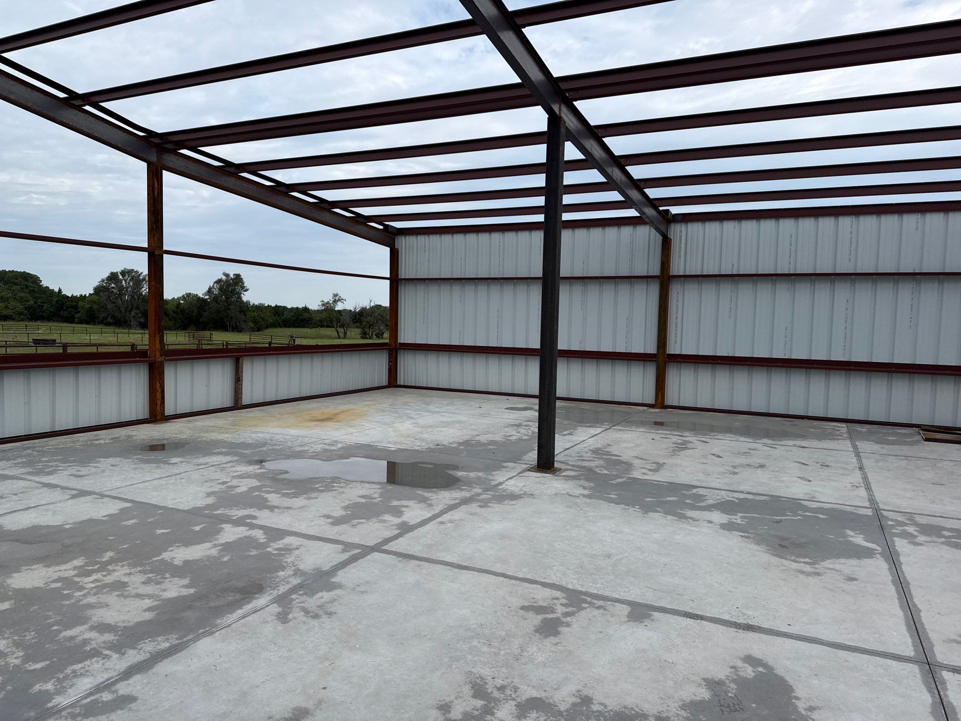 Empty metal framed building, corrugated metal walls, concrete floor, overcast sky visible.