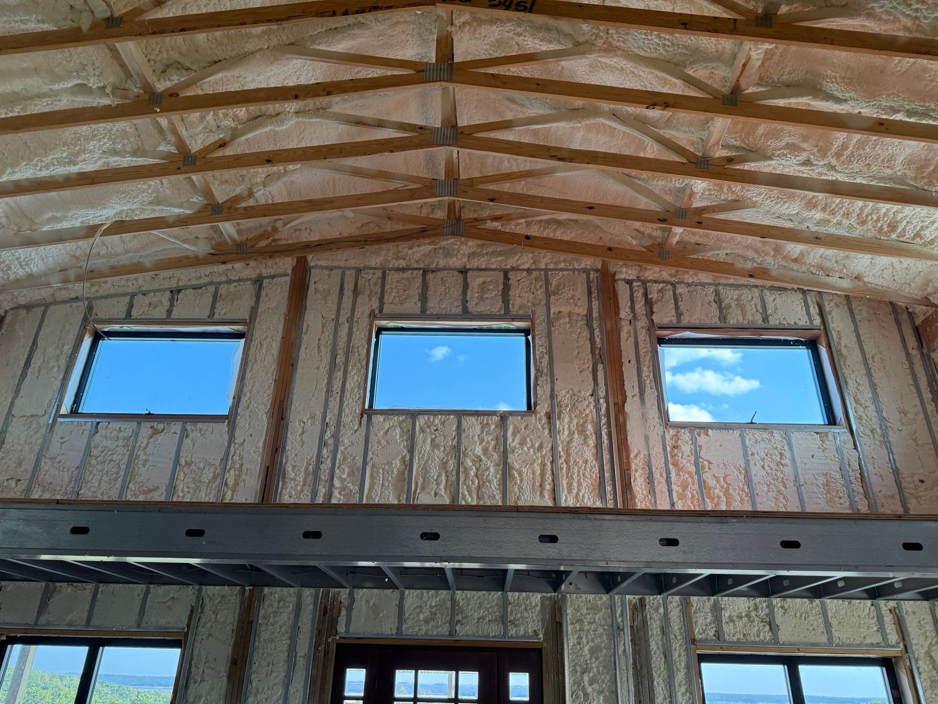Interior view of a building's ceiling and walls. Spray foam insulation covers the wood beams and walls; three windows show blue sky.