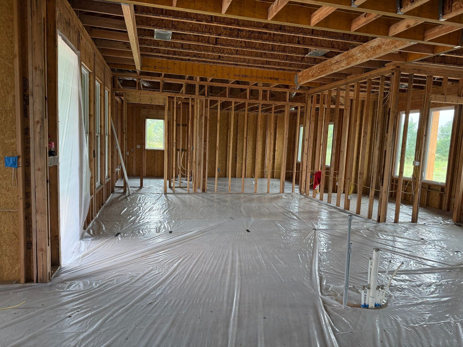 Interior of a house under construction. Wooden framing, plastic sheeting on the floor, and unfinished walls are visible.