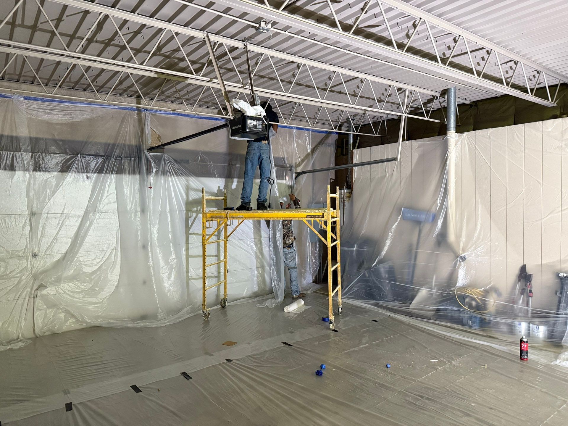 Man on a yellow scaffold working on ceiling infrastructure in a room with plastic sheeting.