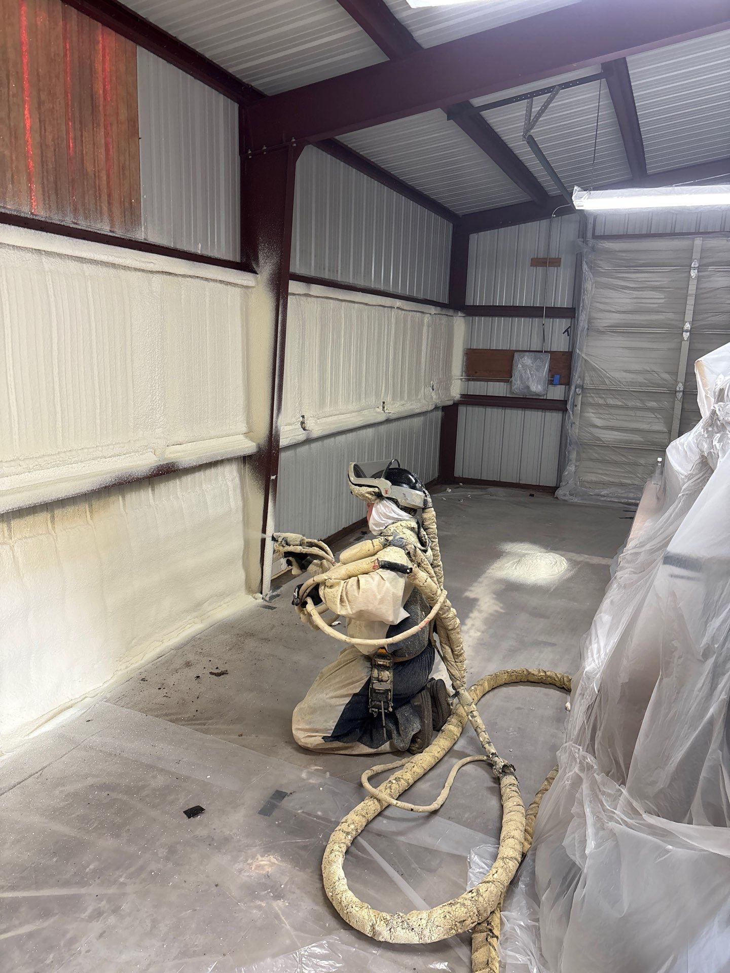 Spray foam insulation being applied inside a metal building with a machine and hose.