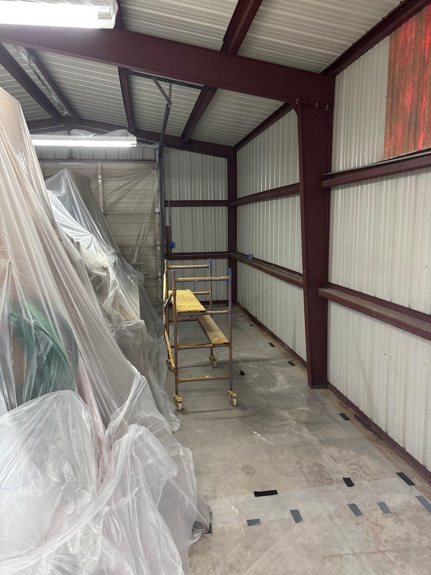 Metal-framed storage building interior. White corrugated walls, burgundy beams, a scaffold, and objects covered in plastic sheeting.