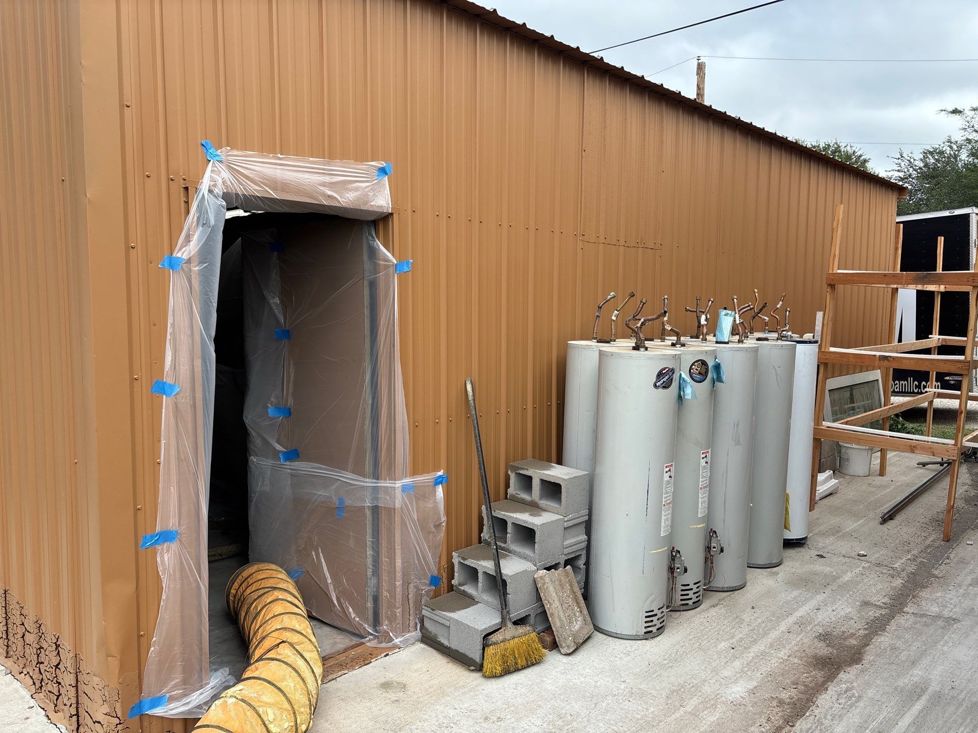 Brown metal building with doorway covered in plastic sheeting. Water heaters and cinder blocks sit outside.