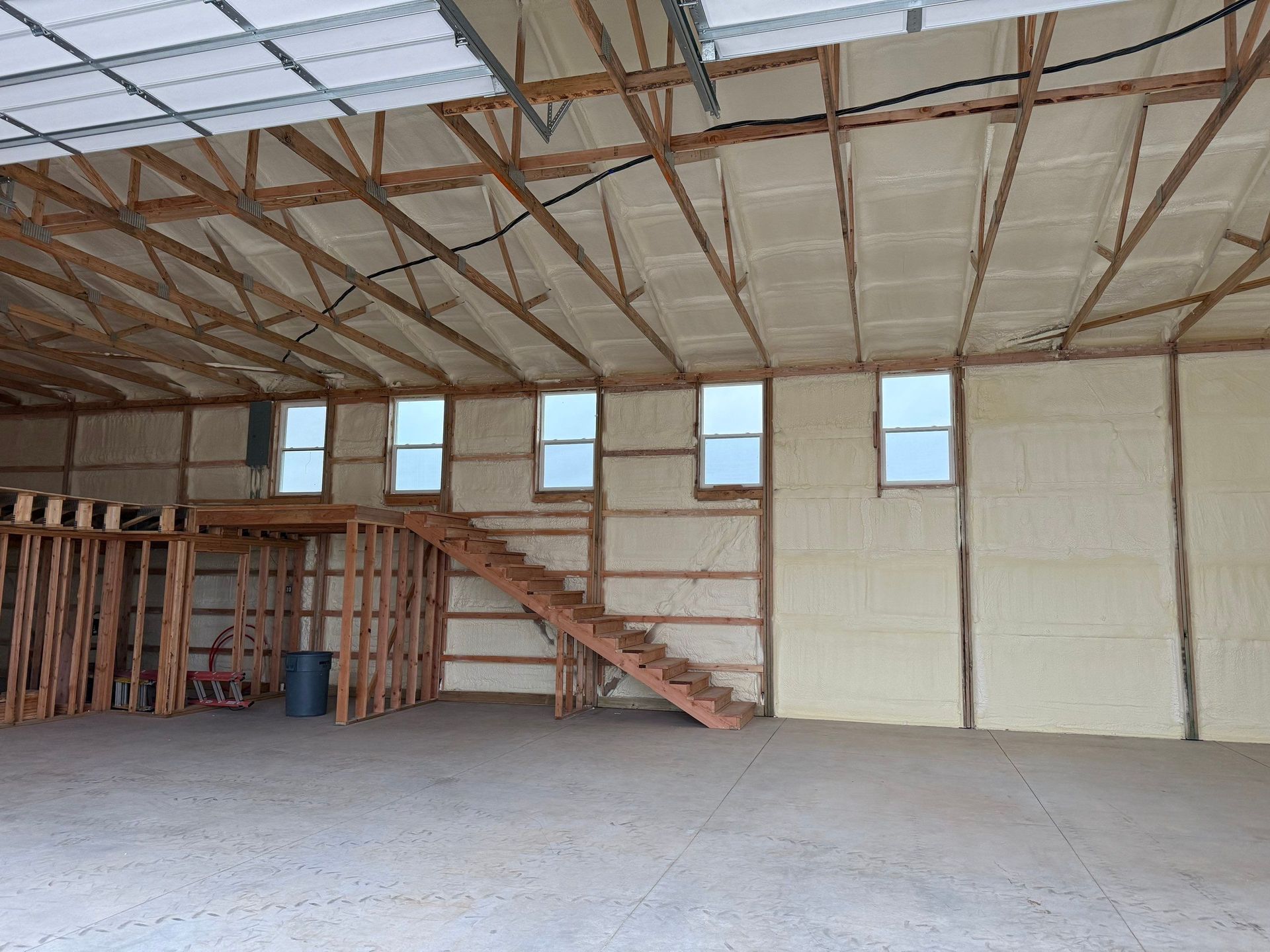 Interior of a large building with exposed wooden beams, foam insulation, windows, and a wooden staircase.
