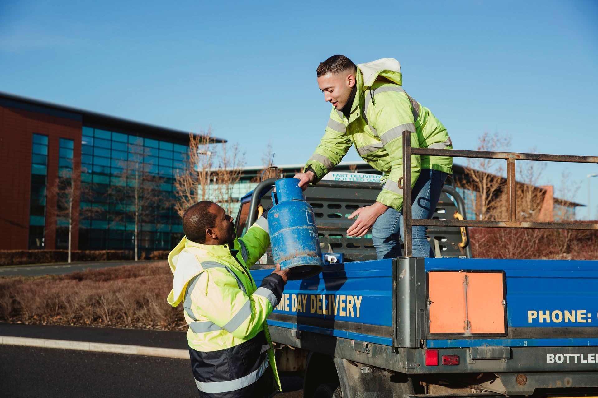 Two workers in hi-vis jackets loading a blue tank onto a trailer outdoors