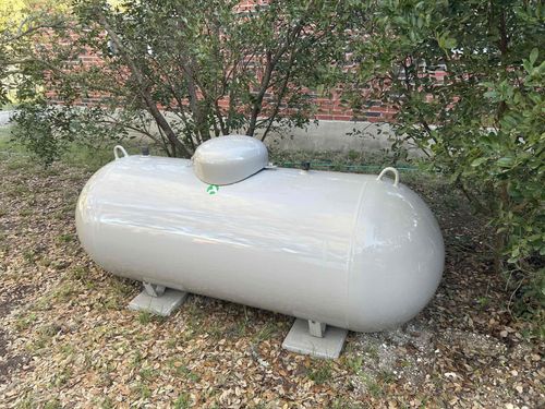 White horizontal propane tank on concrete supports in a leafy outdoor yard