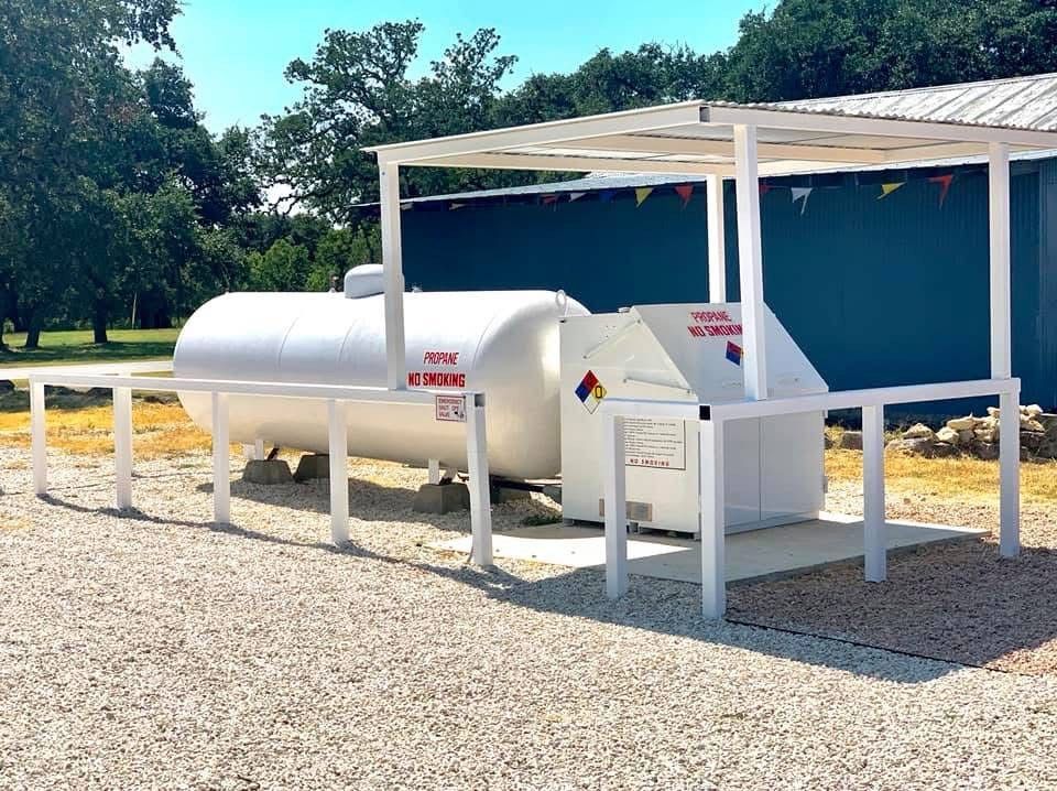 White propane tanks beside a blue utility shelter on a gravel lot, with trees in the background