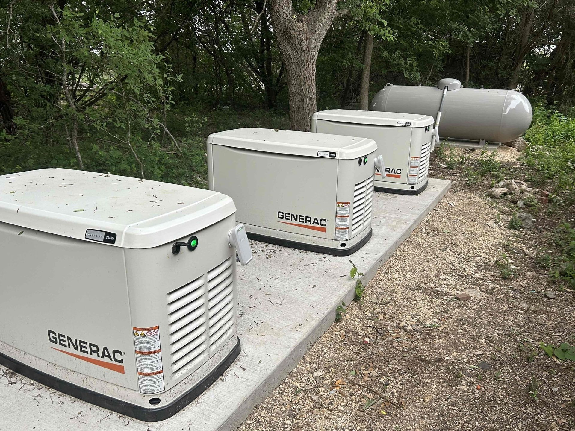 Three white Generac backup generators lined up on a concrete pad beside trees and gravel.