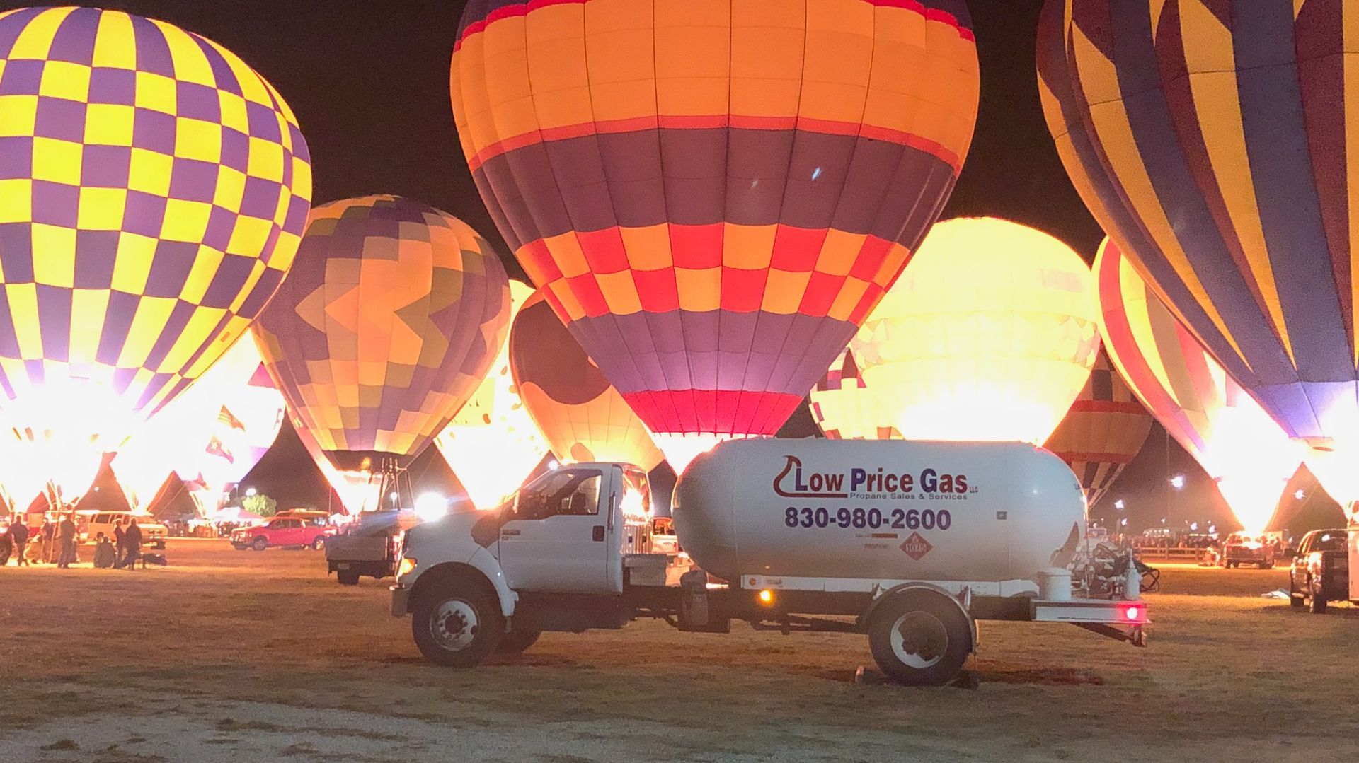Hot air balloons glow at night beside a white truck with a fuel tank in a field.
