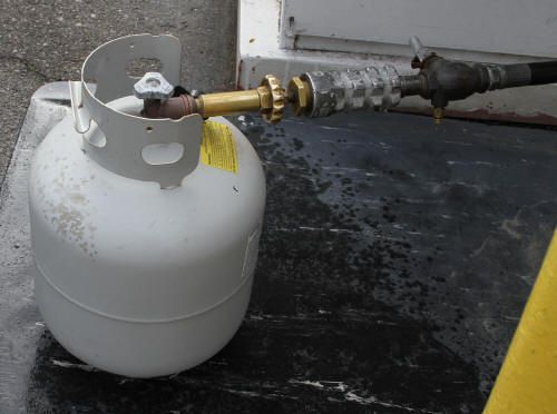 White propane tank with a connected hose on a dark floor, near a wall.