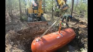 Excavator lifts a large orange tank from a muddy wooded trench using straps.