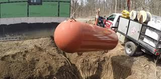 Large orange tank being lifted by crane over a dirt excavation beside a utility trailer.